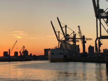 Silhouette cranes at harbor against sky during sunset