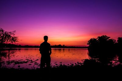 Rear view of silhouette man standing by lake against sky during sunset