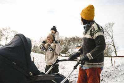 Cheerful woman carrying daughter on shoulder while walking with girlfriend during winter