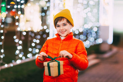 A happy boy in an orange jacket with a gift box in his hands at a christmas market on a winter 