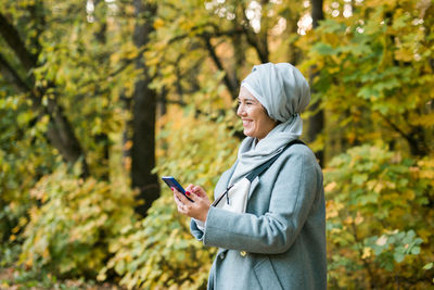 Side view of woman using phone while standing on tree