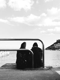 Rear view of friends sitting on jetty while looking at lake against sky