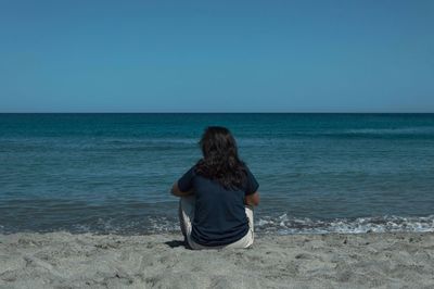 Rear view of woman sitting at beach against clear blue sky