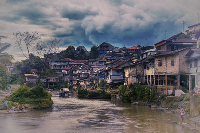 Houses by river amidst buildings in city against sky