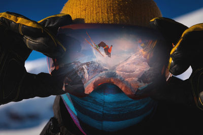 Close-up portrait of a skier in a mask holding on to ski goggles in the reflection of which the