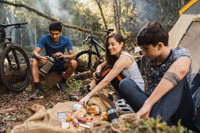 Diverse cyclists sitting on ground near tent and having picnic while hiking together in forest