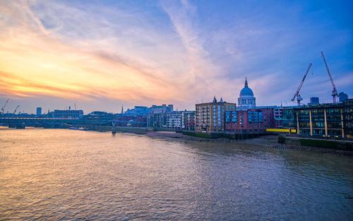 Bridge over river by buildings against sky during sunset