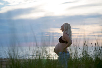 Pregnant woman standing amidst grass at beach against cloudy sky