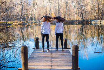 Full length of friends tossing hair while standing on pier by lake in forest