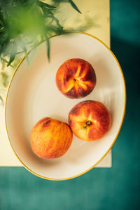 Three peaches in bowl next to vase of tarragon on golden table, teal