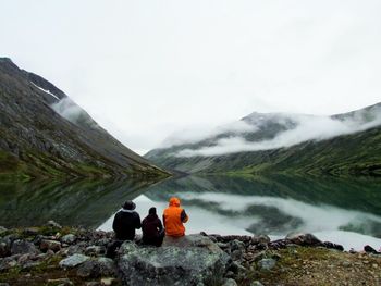 Rear view of people sitting on mountain by lake against sky