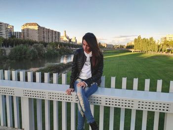 Young woman sitting on retaining wall at park in city against sky