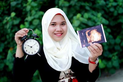 Portrait of young woman holding alarm clock with ultrasound standing against plants