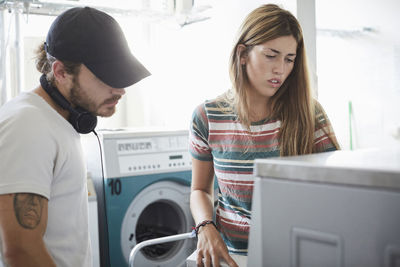 Man assisting female friend in using washing machine at laundromat during sunny day