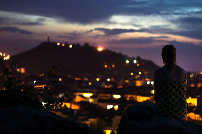 Rear view of woman sitting against illuminated city during sunset