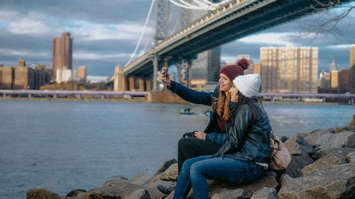 Woman sitting on rock by sea against sky in city