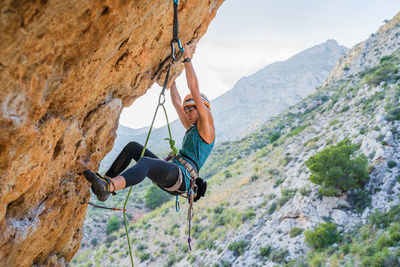 From below side view of active focused youthful female alpinist climbing on cliff in summer day