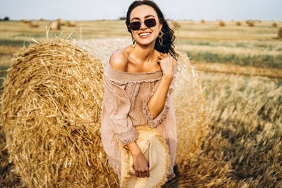 Smiling woman in sunglasses with bare shoulders on a background of wheat field and bales of hay.