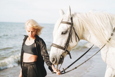 Young blonde woman in black clothes riding white horse on seascape background