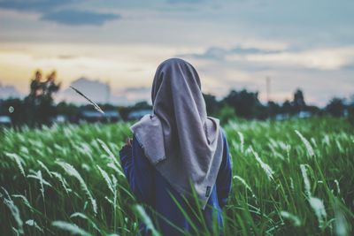 Rear view of young woman wearing hijab standing amidst plants against sky in farm during sunset