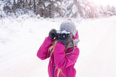 Backlit portrait of girl in warm clothes taking photo in winter forest. 