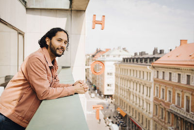 Smiling man looking away while leaning on hotel balcony during sunny day