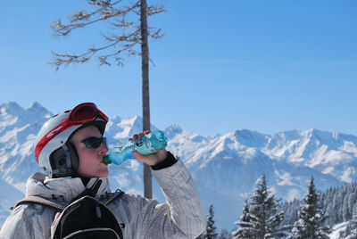 Woman wearing skiing helmet drinking water against snowcapped mountain