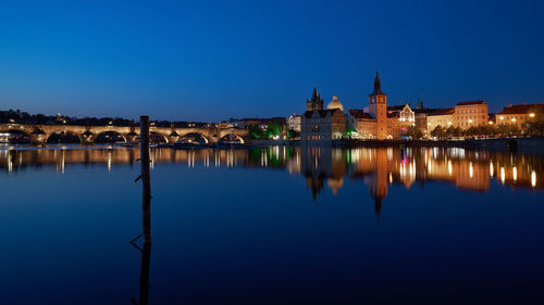 Bridge over river by illuminated town against clear blue sky