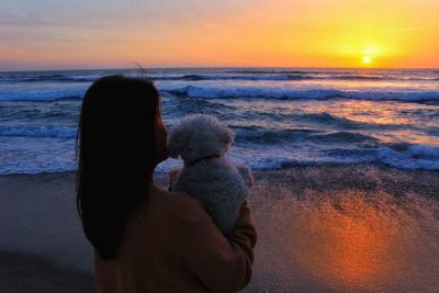 Rear view of woman carrying dog while looking at sea during sunset