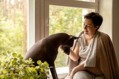 Middle age beautiful woman sitting on windowsill with her dog. 50-year-old woman