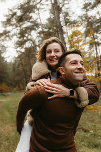 Couple having fun in the autumn park outdoors, happy man is carrying a woman on his back in woods