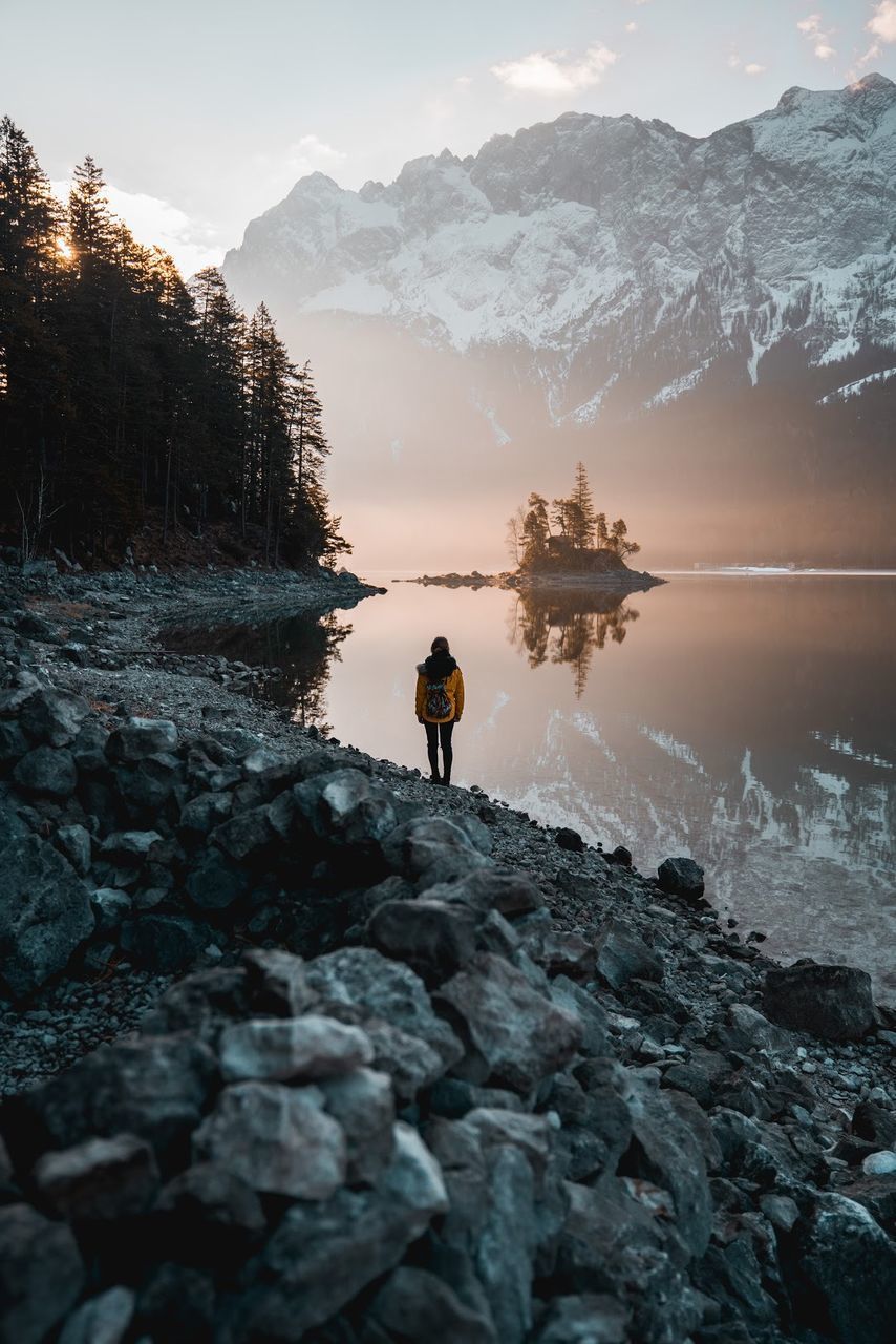 REAR VIEW OF MAN STANDING ON ROCK BY LAKE AGAINST SKY