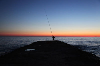 Silhouette man fishing in sea against sunset sky