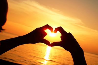 Cropped image of person making heart shape against sea during sunset