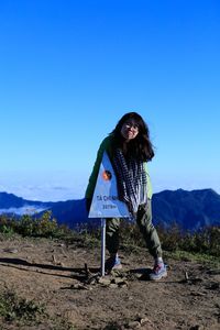 Portrait of woman making face while standing by road sign on mountain peak against blue sky