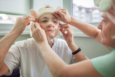 Two happy senior women apply facial cosmetic mask, have fun at home, body positivity, ageism