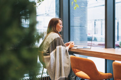 Adult smiling brunette woman in grey warm scarf working with laptop on table at cafe in cold season