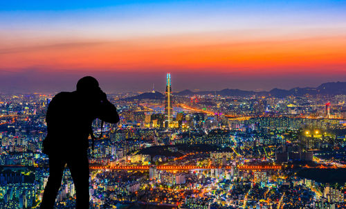Rear view of silhouette man photographing cityscape against sky during sunset