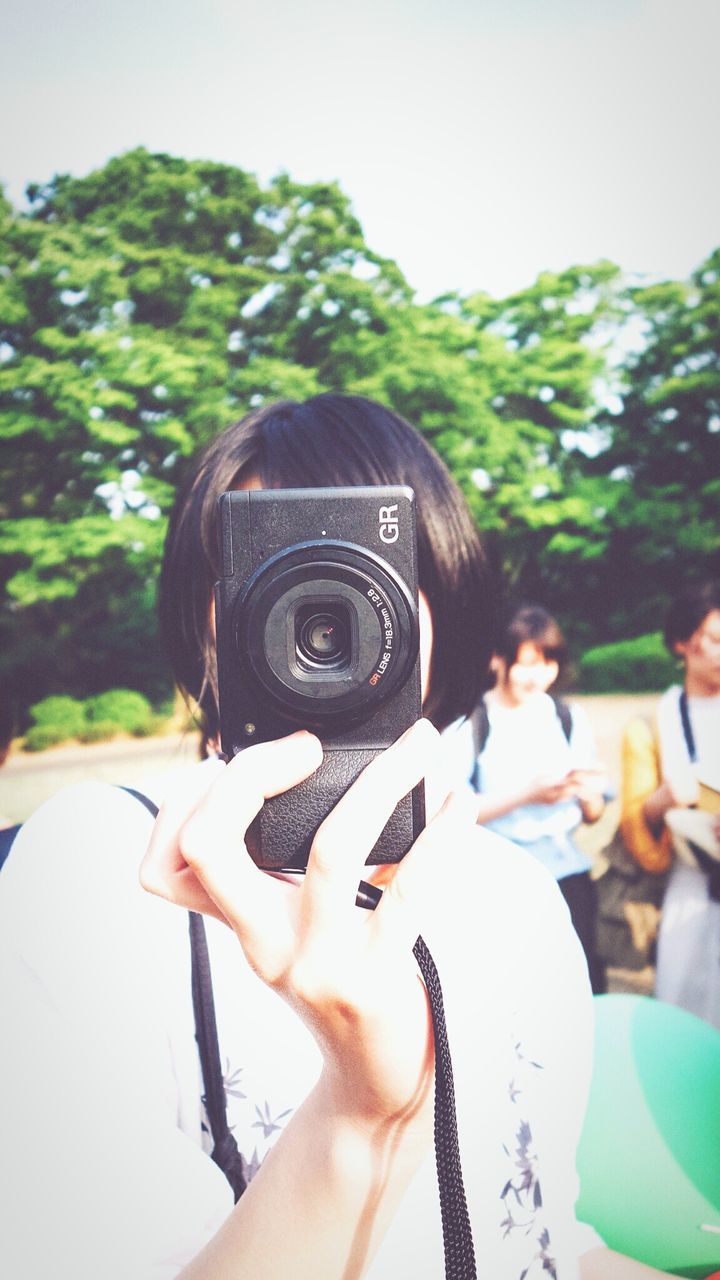 real people, photography themes, photographing, camera - photographic equipment, technology, one person, leisure activity, holding, digital camera, day, tree, focus on foreground, headshot, childhood, outdoors, lifestyles, photographer, digital single-lens reflex camera, women, close-up, men, human hand, sky, people