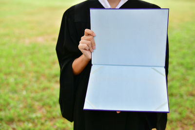 Midsection of woman in graduation gown holding blank folder while standing on field