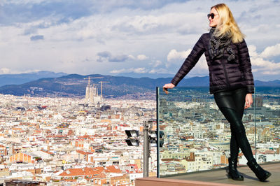 Young woman wearing warm clothing while standing on balcony against cityscape