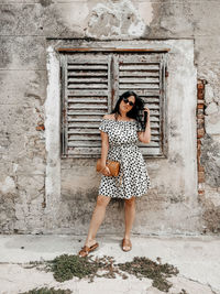 Front view of young woman in polka dot dress posing in front of weathered wall and old window.