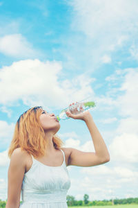 Young woman drinking water from bottle against cloudy sky