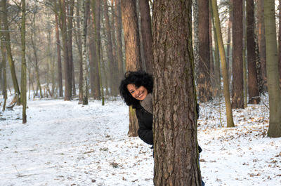 Portrait of cheerful woman standing behind tree on snow covered field