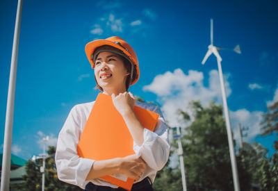 Female engineer in hardhat holding file while standing against windmill