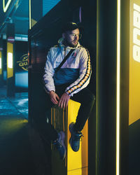 Young man looking away while standing against illuminated yellow wall