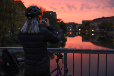 Man photographing bicycle on railing against sky during sunset