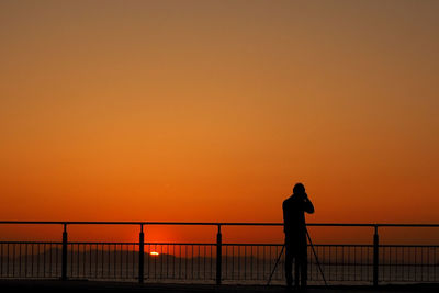 Silhouette man standing by railing towards sea against clear orange sky