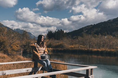 Young woman sitting on fence on shore of lake in autumn