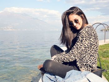 Smiling young woman sitting by sea against sky